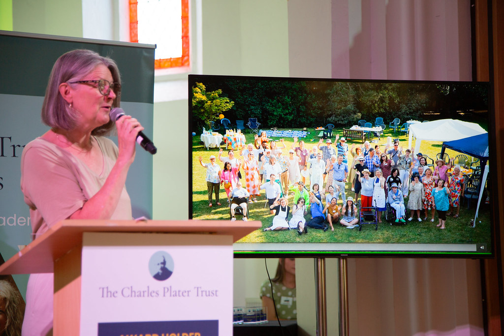 A person speaking into a mic in front of a screen showing Lyn's House people